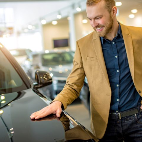 Homem sorridente admira seu novo carro em uma concessionária, simbolizando a realização de um sonho conquistado com planejamento financeiro por meio do consórcio automotivo, sem entrada e sem juros.