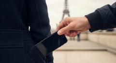 Foto em close-up extremo mostrando o momento exato de um furto em Paris. A mão de um ladrão retira um smartphone do bolso de trás de um turista, com o aparelho já no meio do caminho para fora. A cena é focada na ação, sem mostrar rostos, e tem a Torre Eiffel desfocada ao fundo para ilustrar o risco em viagens internacionais.
