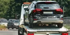 Um carro preto com a traseira danificada está sendo transportado por um caminhão guincho em uma estrada movimentada. A imagem representa o serviço de assistência 24 horas do seguro auto Pattini, que oferece suporte imediato como reboque, socorro mecânico, troca de pneus e mais, em qualquer hora do dia.