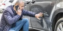 Homem agachado ao lado do carro danificado, observando arranhões na porta enquanto fala ao celular, demonstrando preocupação. A imagem representa a cobertura de colisões do seguro auto Pattini, que oferece suporte e indenização em casos de batidas ou acidentes.