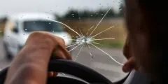 Vista do interior de um carro com foco no para-brisa rachado por um impacto. A mão do motorista está no volante, e a estrada aparece ao fundo. A imagem ilustra a cobertura de danos a vidros do seguro auto Pattini, que também pode incluir proteção para faróis, lanternas e retrovisores em caso de trincas, estilhaços ou quebras acidentais.