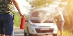 Um homem se aproxima de um carro com o capô aberto e fumaça saindo do motor, segurando um extintor de incêndio. A cena mostra um princípio de incêndio em via pública, ilustrando a cobertura contra incêndios do seguro auto Pattini, que protege o veículo em situações de fogo acidental, explosões ou raios.