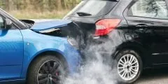 Dois carros colidiram em uma via: um veículo azul teve a dianteira danificada ao bater na traseira de um carro preto, com fumaça saindo do local do impacto. A imagem ilustra a cobertura de danos a terceiros do seguro auto Pattini, que protege o segurado em caso de prejuízos causados a outros veículos, pessoas ou propriedades.