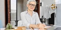 Mulher com cabelos grisalhos e expressão serena sentada à mesa, simbolizando a importância da cobertura de danos morais ou estéticos que podem ser causados por atividades empresariais.