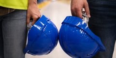 Duas pessoas segurando capacetes de segurança azul, representando a proteção contra morte ou acidente de funcionários sob responsabilidade do empregador em ambientes de risco.