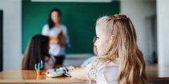 Criança em sala de aula observando a professora ao fundo, simbolizando o amparo financeiro para educação de dependentes garantido em coberturas especiais do Seguro de Vida Empresarial.